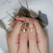 Trendy beige almond nails featuring animal spots and gold embellishments